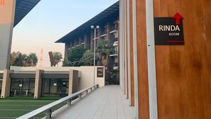 A corridor with a sign for 'RINDA ROOM' and a 'RINDA Fitness' sign, leading to a building with a modern design and a sports field visible in the background. - Phurinda Residence (Nong Khae)