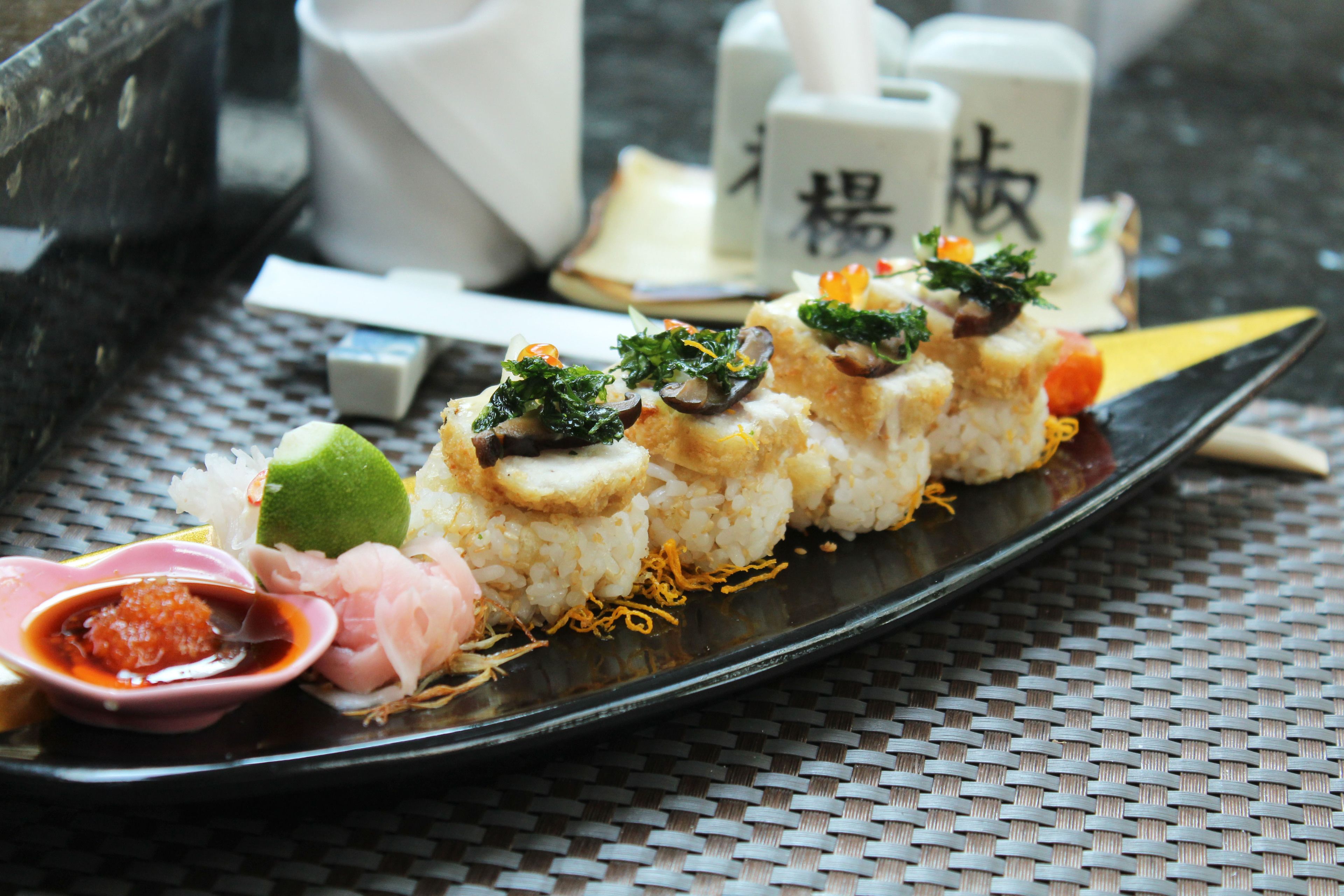 Sushi-lautanen, jossa on erilaisia koristeita, ja pieni kulho kastiketta mustalla lautasella.