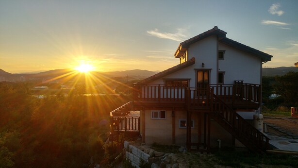 Ein zweistöckiges Haus mit Balkon, Treppe und Blick auf einen Sonnenuntergang über einer Berglandschaft.