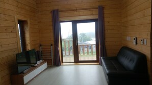 Ein Zimmer mit Holzwandverkleidung, einem Fernseher, einem schwarzen Ledersofa, einer Schiebetür zu einem Balkon und einem kleinen Tisch mit einem Fernseher-Schrank.