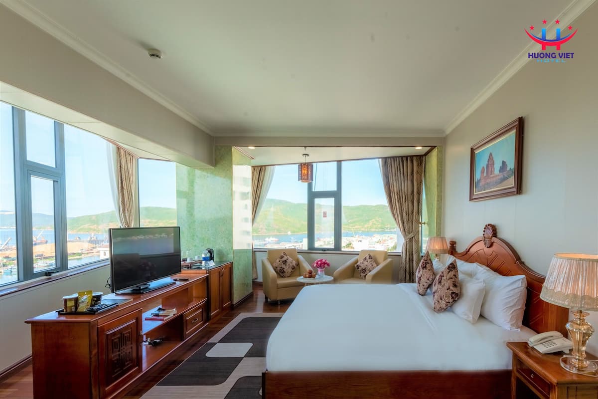A hotel room with a large bed, a television, a desk with a computer, and a view of the beach.