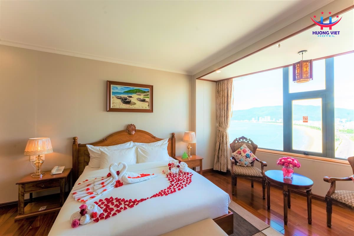 A hotel room with a bed, a nightstand with a lamp, a chair, a small table with a vase, and a window with a view.