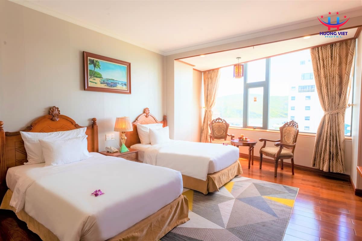 A hotel room with two beds, a painting, a window with curtains, a small table, and a chair.