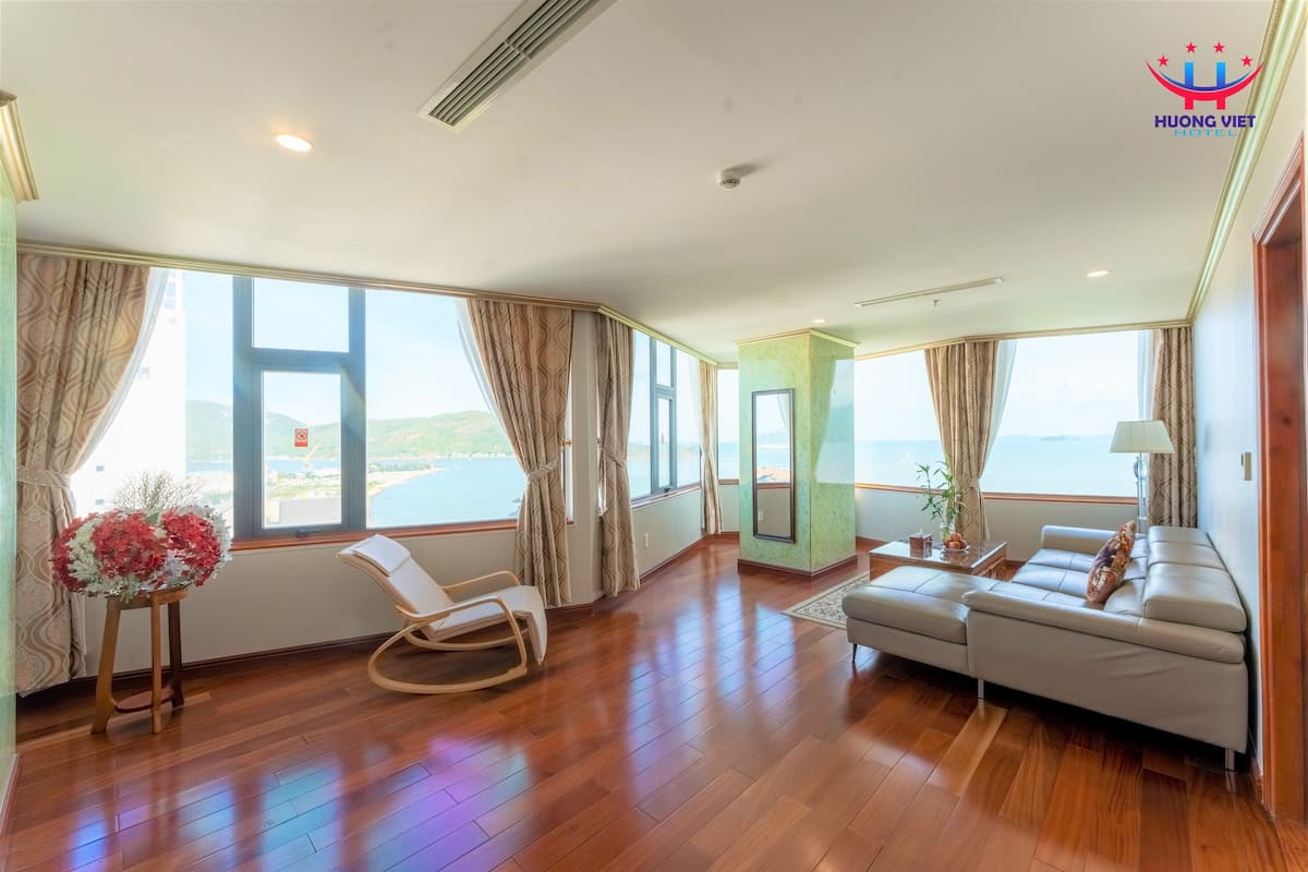 A spacious room with wooden flooring, a white sofa, a rocking chair, and large windows offering a view of the sea.