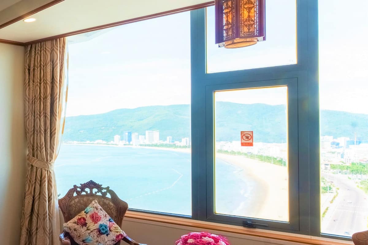 A room with a large window overlooking a coastal view, a table with a vase of flowers, and two upholstered chairs.