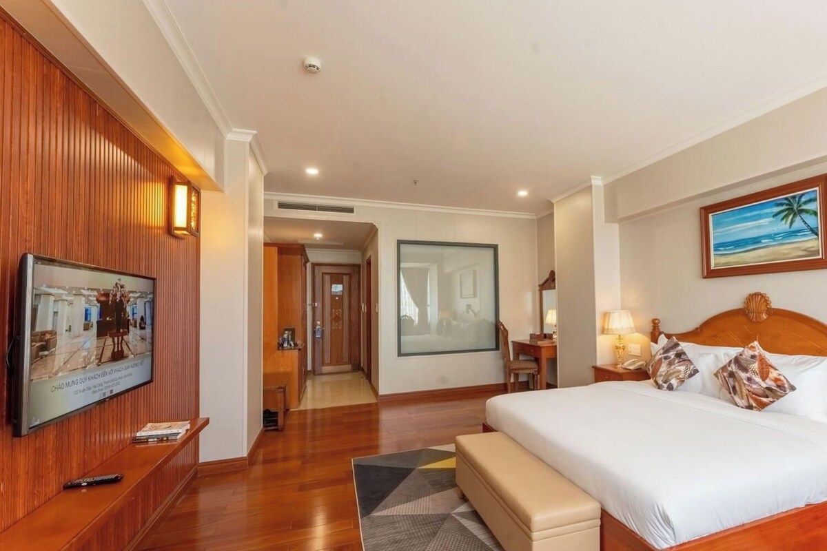 A hotel room with a large bed, a television mounted on the wall, a desk with a chair, and a painting on the wall.