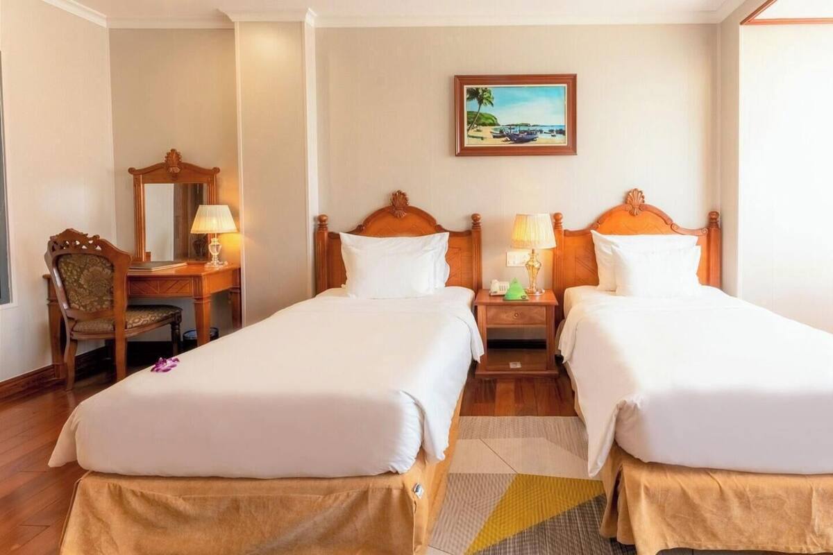 A hotel room with two beds, a desk, a chair, a nightstand, a lamp, a painting, and a window with curtains.