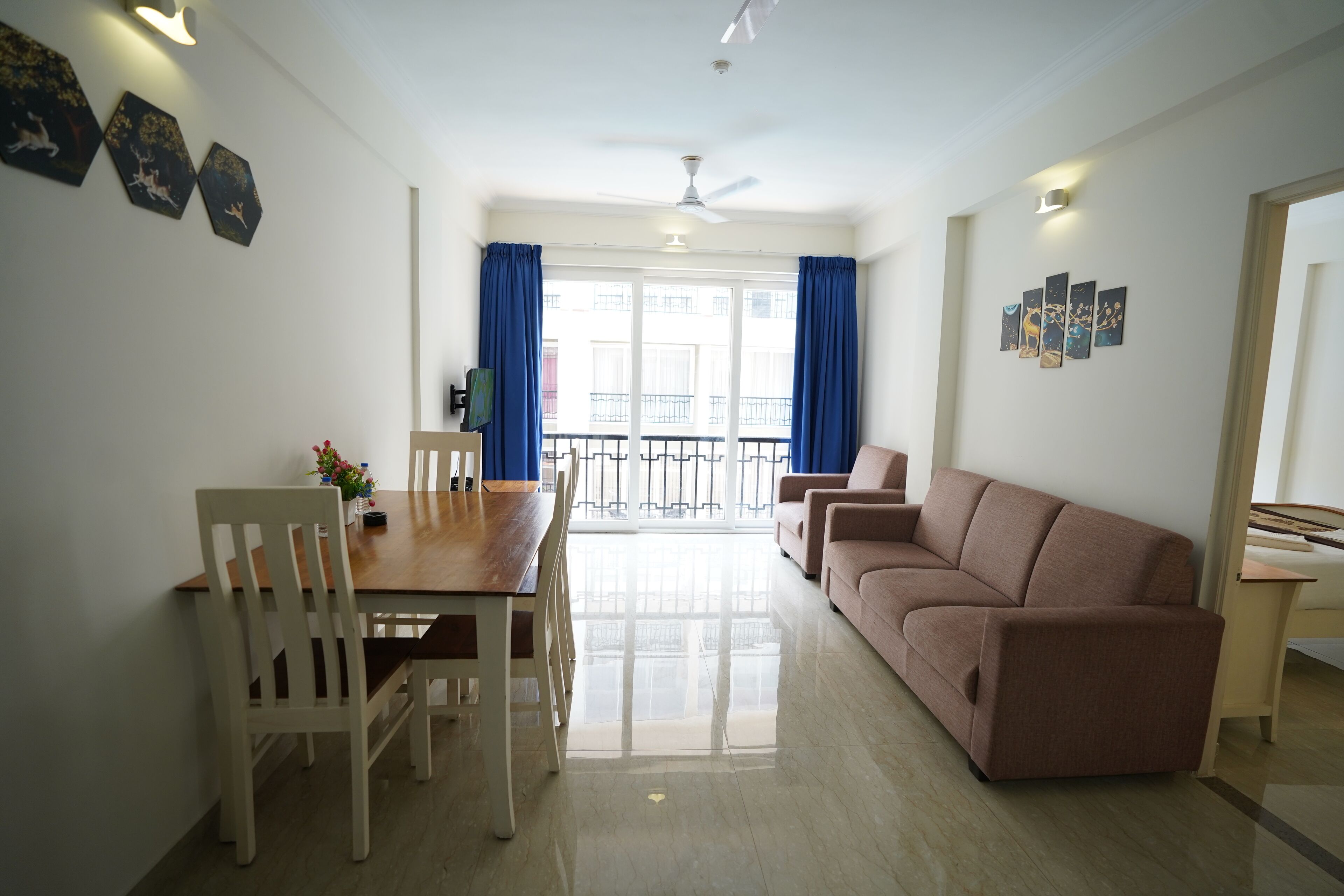 A spacious living area with a wooden dining table, white chairs, a beige sofa, and a balcony with blue curtains.