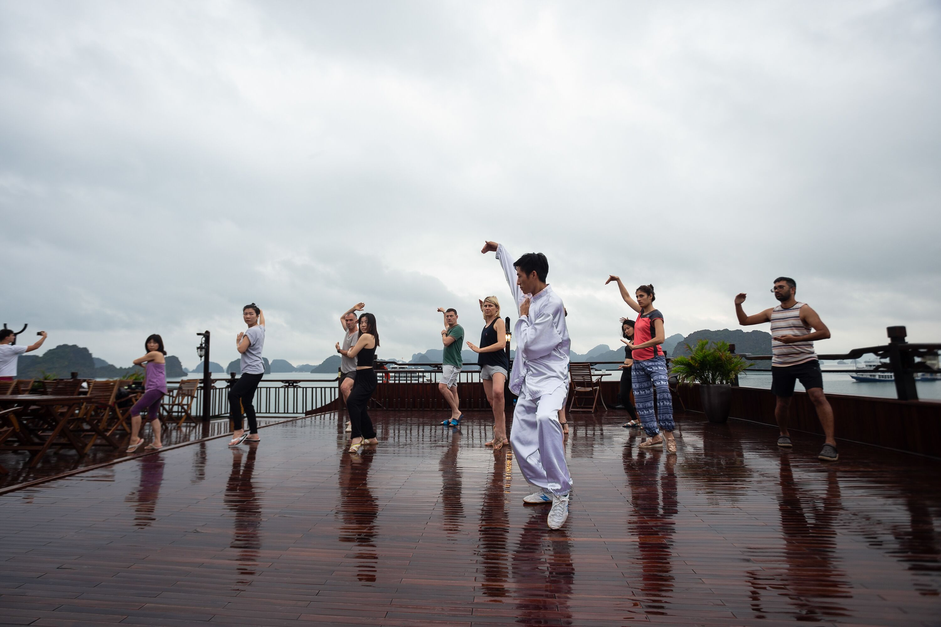A group of people performing a dance routine on a wooden deck with a view of a body of water and mountains in the background.