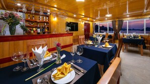 A restaurant with wooden interiors, tables set with yellow napkins, wine glasses, and a fruit basket. - Azela Cruise (Hai Phong)