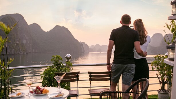 A couple overlooking a body of water with mountains in the background, dining table with food and drinks, and outdoor seating. - Azela Cruise (Hai Phong)