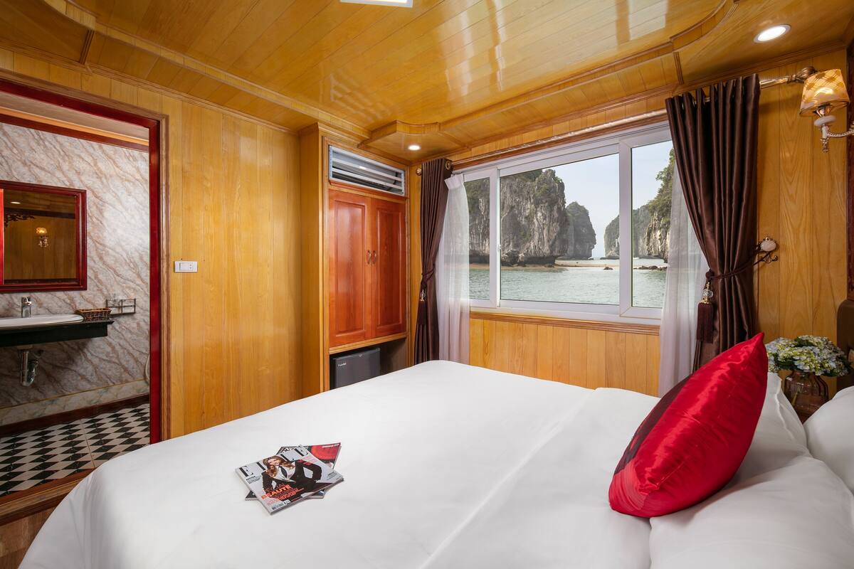 A hotel room with a wooden interior, a bed with a red pillow, a window with curtains, and a view of a coastal landscape.