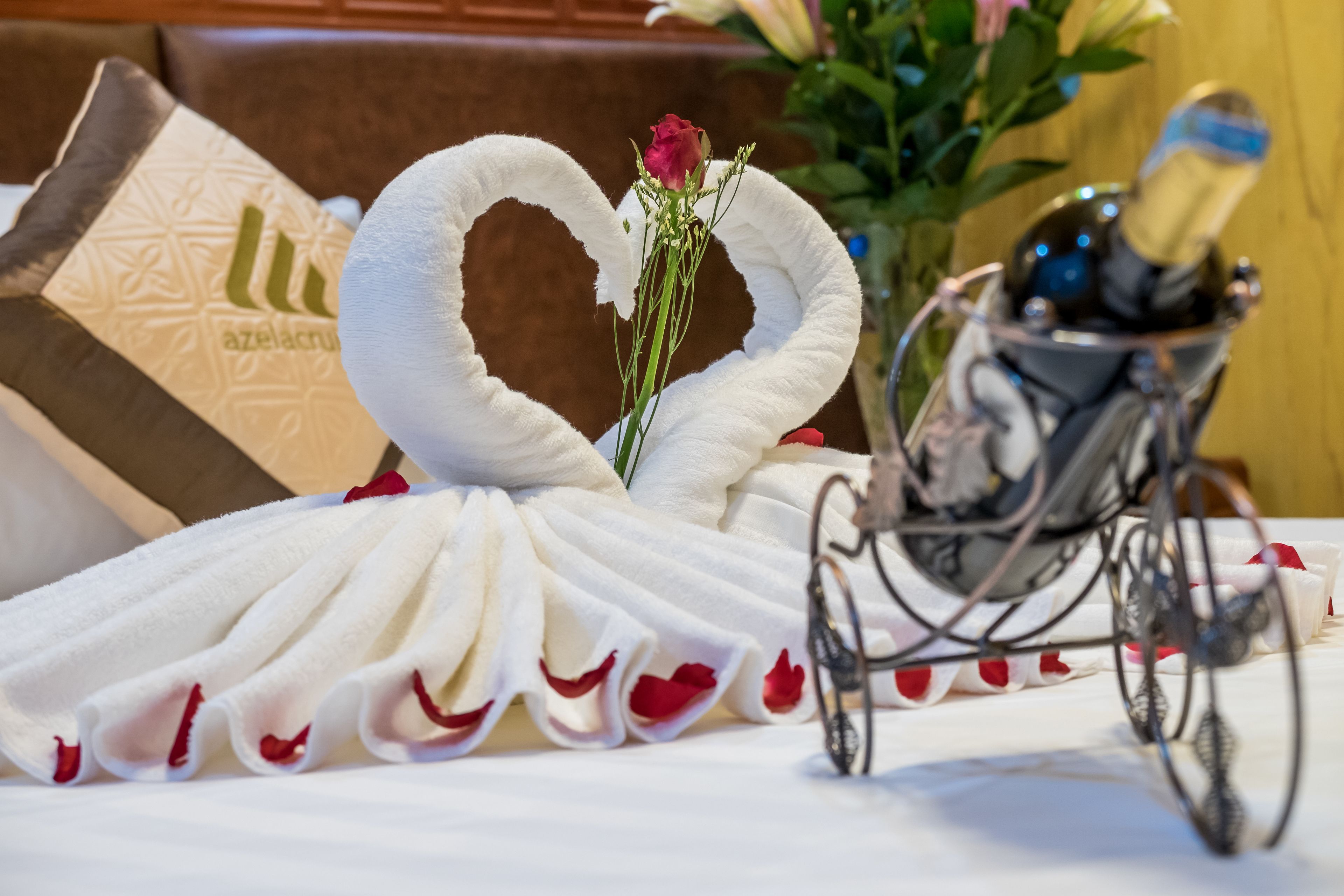 A table with a white towel arranged in a heart shape, a rose, and a champagne bottle in a glass holder.