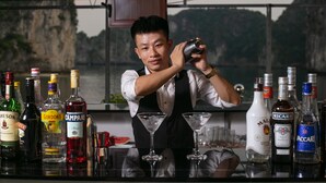 A bartender preparing a drink at a bar with various bottles of alcohol and empty glasses on the counter. - Azela Cruise (Hai Phong)