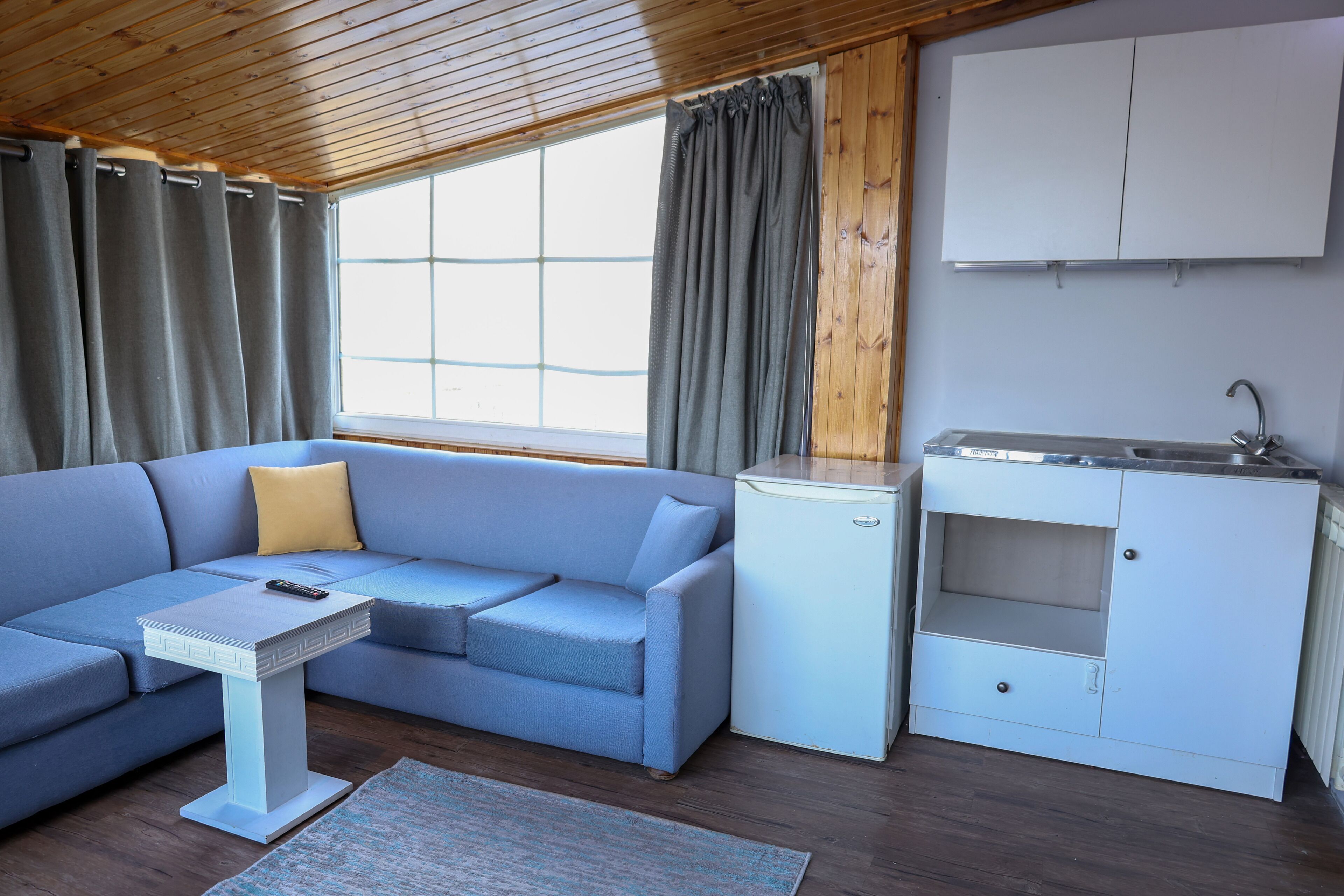 A modern, compact living space with a sofa, a small table, a kitchenette, and a window with curtains.