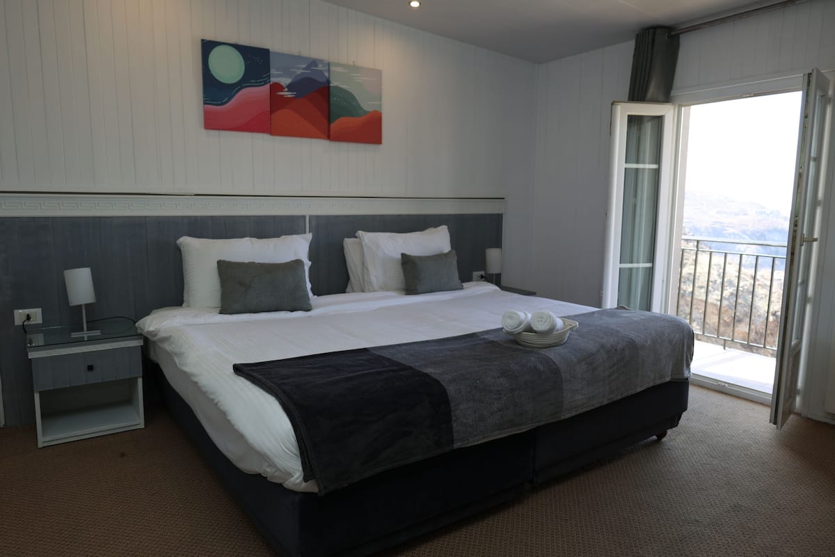 Une chambre d’hôtel moderne avec un grand lit, une table de chevet, une lampe et une vue sur les montagnes par la fenêtre.