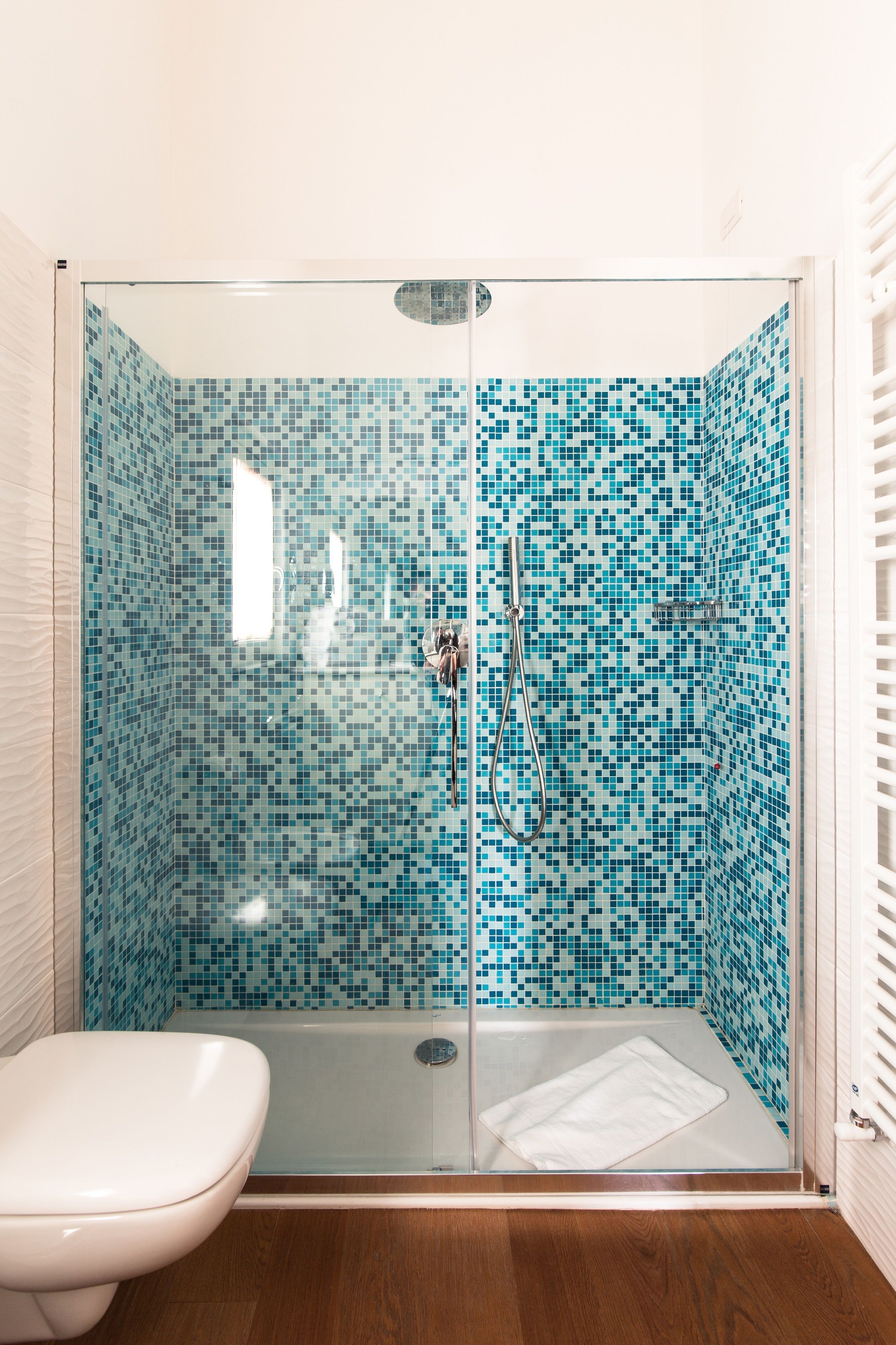 Chuveiro com divisória de vidro, paredes com azulejos em mosaico, vaso sanitário branco e soalho de madeira.
