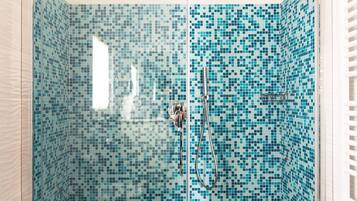 Chuveiro com divisória de vidro, paredes com azulejos em mosaico, vaso sanitário branco e soalho de madeira.