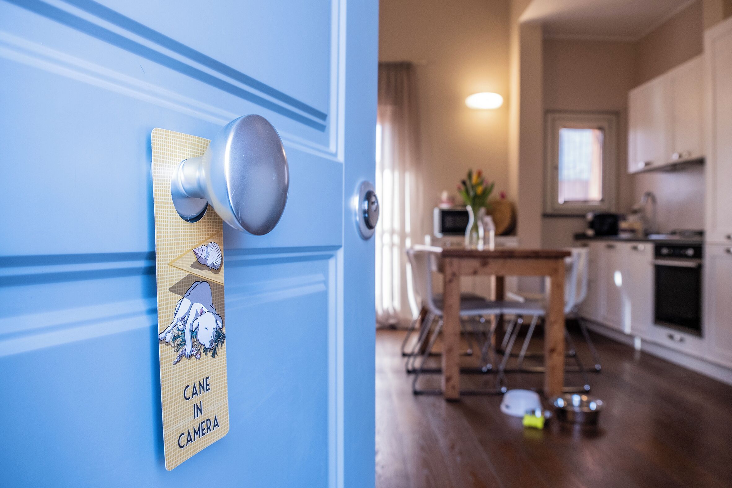 Una puerta con un cartel que dice "CANE IN CAMERA", una mesa de comedor de madera y una cocina con armarios y electrodomésticos.