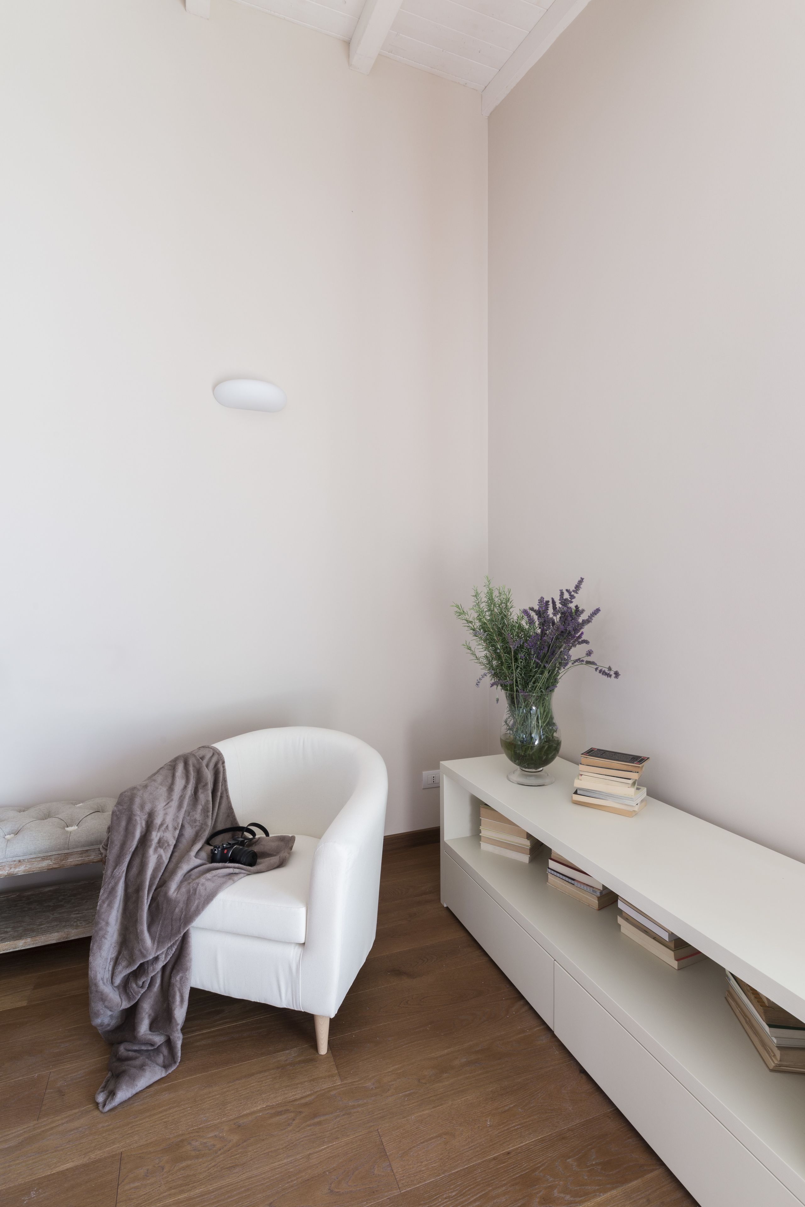 Una habitación minimalista con un sillón blanco, una consola blanca con un jarrón de flores y una pila de libros.