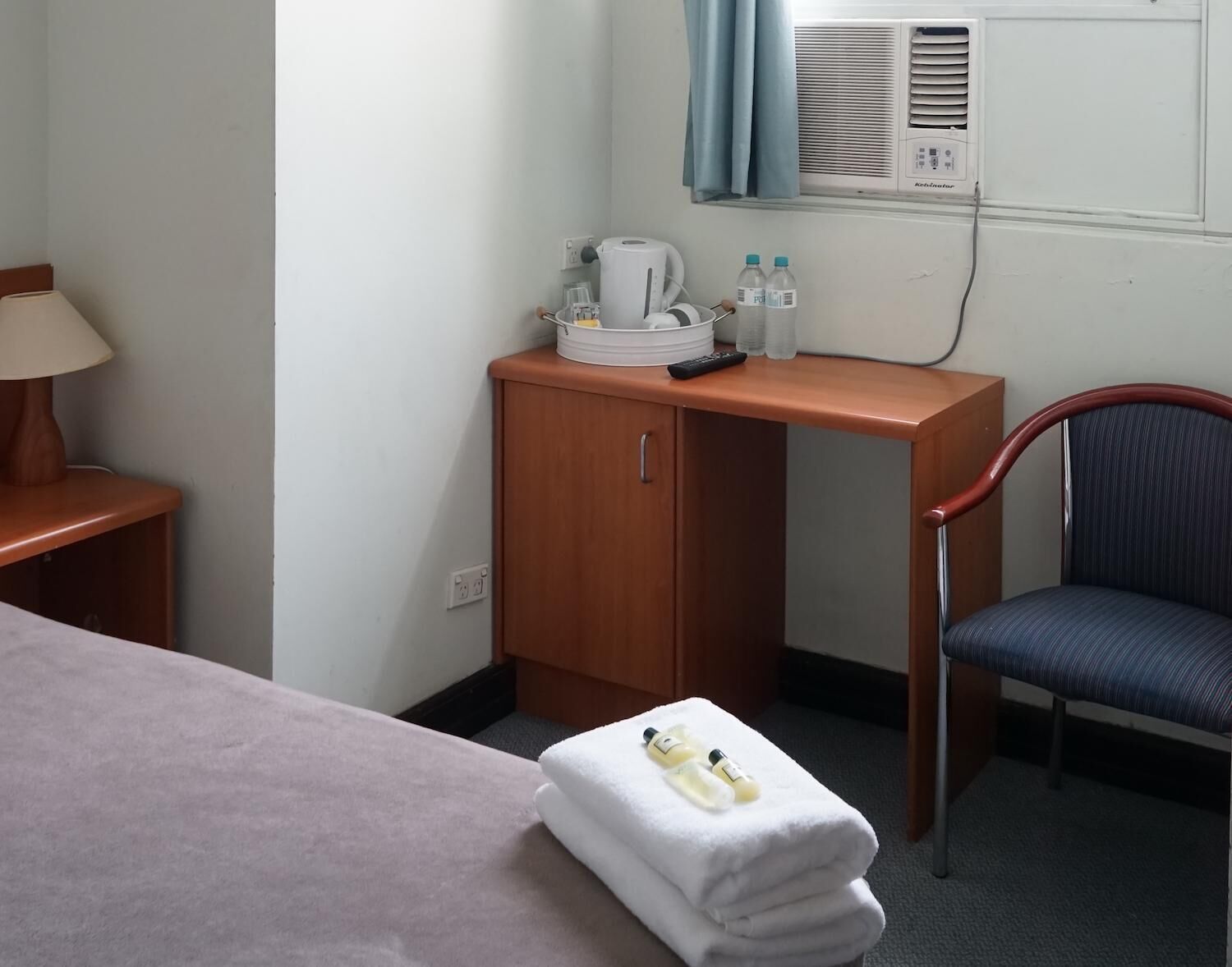 A hotel room with a bed, a wooden desk with a kettle, a chair, and a small table with toiletries.