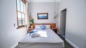 Una cama bien hecha con una colcha blanca, dos almohadas, una mesita de noche con una lámpara y un cuadro en la pared.