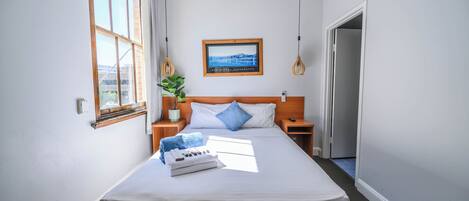 A neatly made bed with a white comforter, two pillows, a small bedside table with a lamp, and a framed picture on the wall.