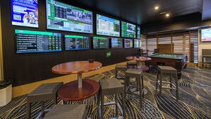Un área de casino con varias pantallas mostrando información sobre apuestas deportivas, mesas de billar y asientos cómodos.