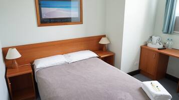 Una cama bien hecha con cabecera de madera, dos lámparas de noche, una mesita de noche de madera, un escritorio con lámpara, una cafetera y una ventana con cortinas.