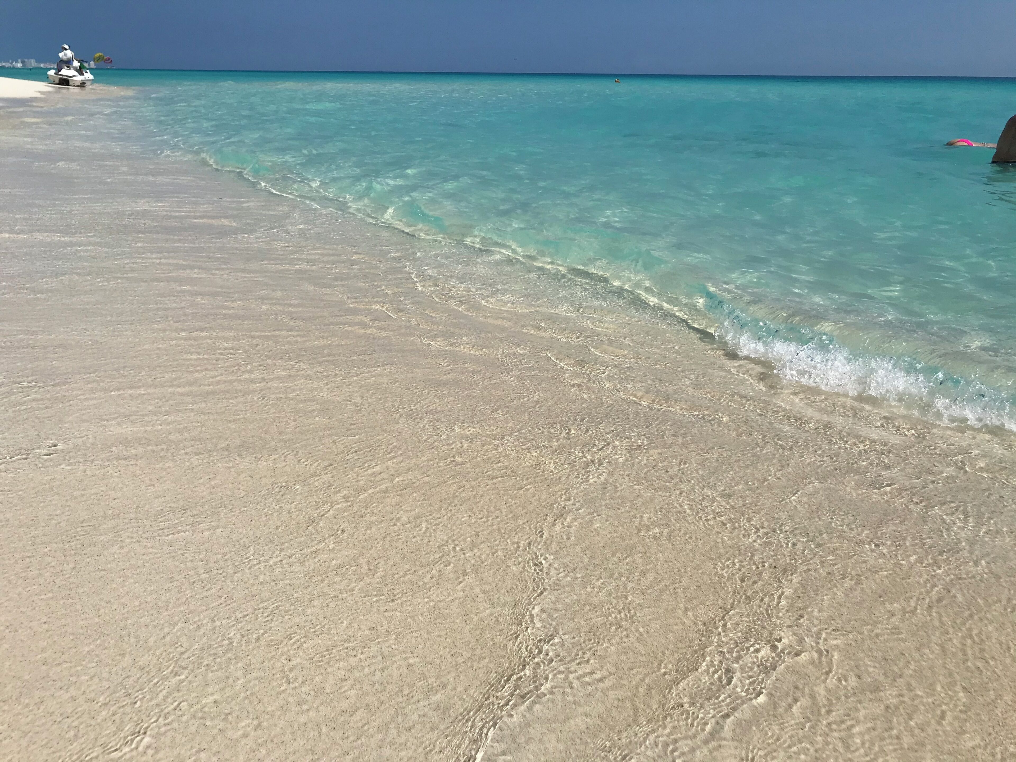 A beach with clear turquoise water, a sandy shore, and a jet ski in the distance.