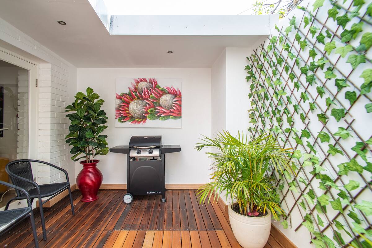 A patio with a grill, a chair, a potted plant, and a wall art piece.