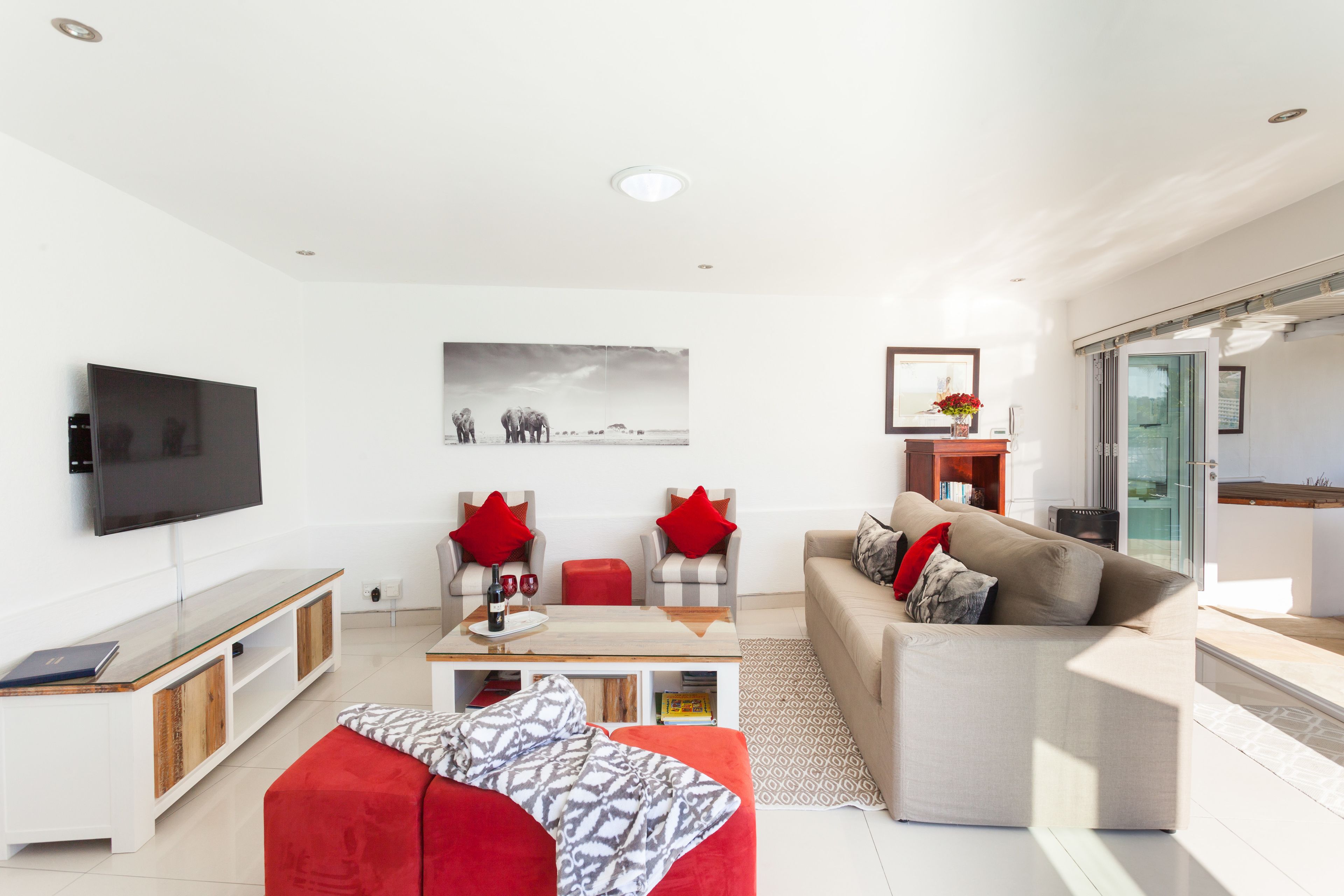 A modern living room with a flat-screen TV, a sofa, a red ottoman, and a coffee table.