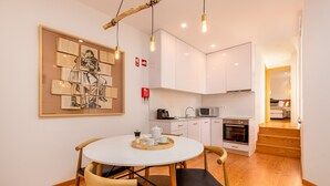 Een moderne keuken met witte kasten, een magnetron en een oven. Er is een eethoek met een ronde tafel en stoelen, en een kunstwerk in een lijst aan de muur.