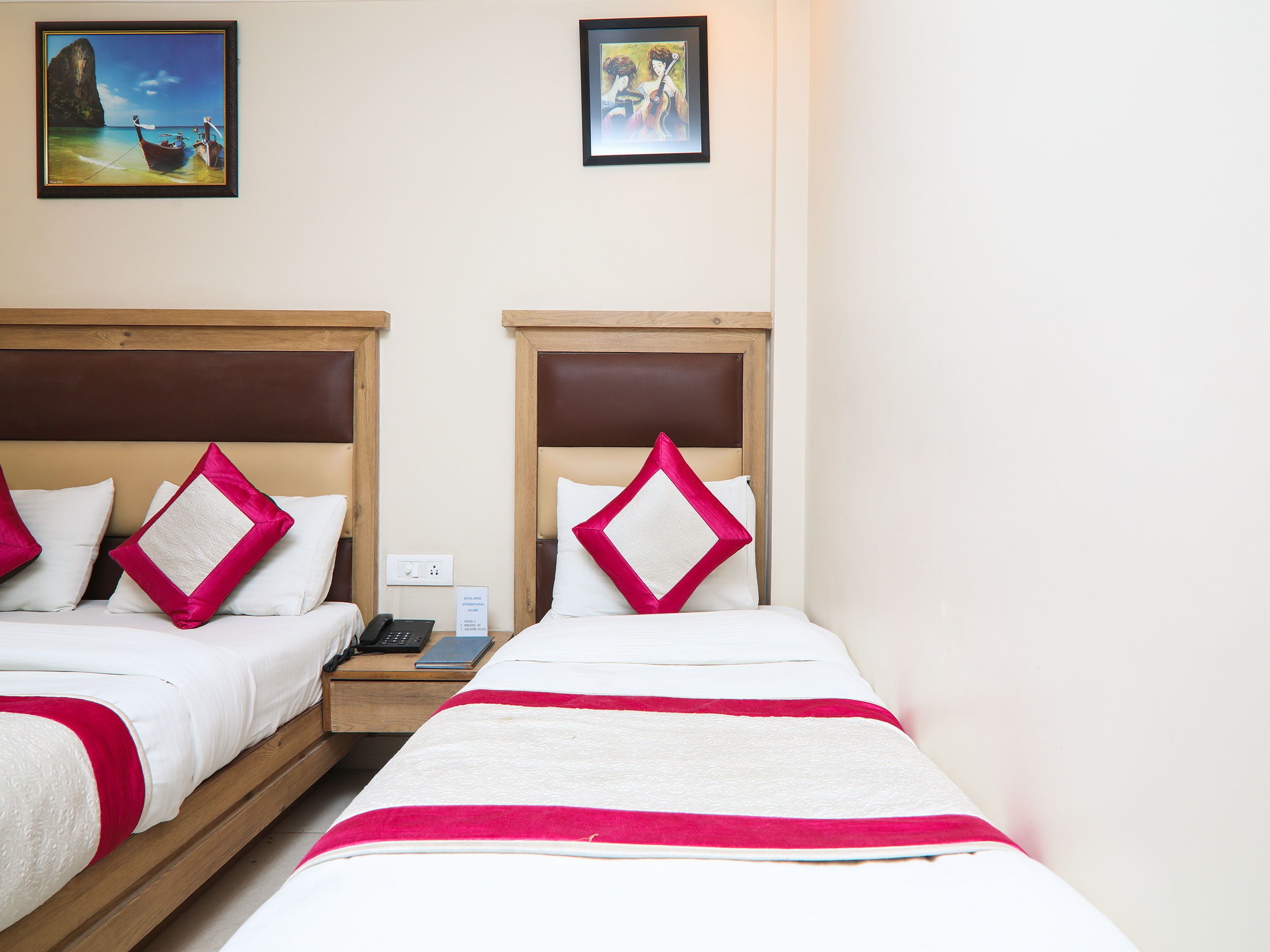 A hotel room with two beds, each with a wooden headboard, white bedding, and red and white pillows. There are two framed pictures on the wall above the beds.