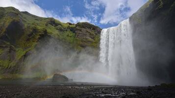 Une cascade surmontée d’un arc-en-ciel, entourée de collines verdoyantes.
