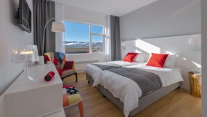 A modern hotel room with two beds, a desk, a chair, a TV, and a large window with a view of mountains. - Hotel Selja (Rangárþing eystra)