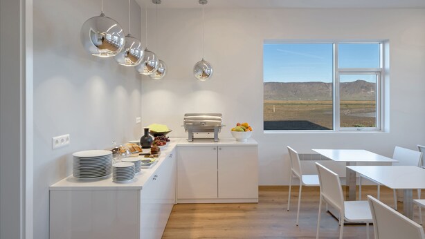 A modern kitchen with white cabinetry, a buffet counter with plates and fruits, and a view of the desert through large windows. - Hotel Selja (Rangárþing eystra)