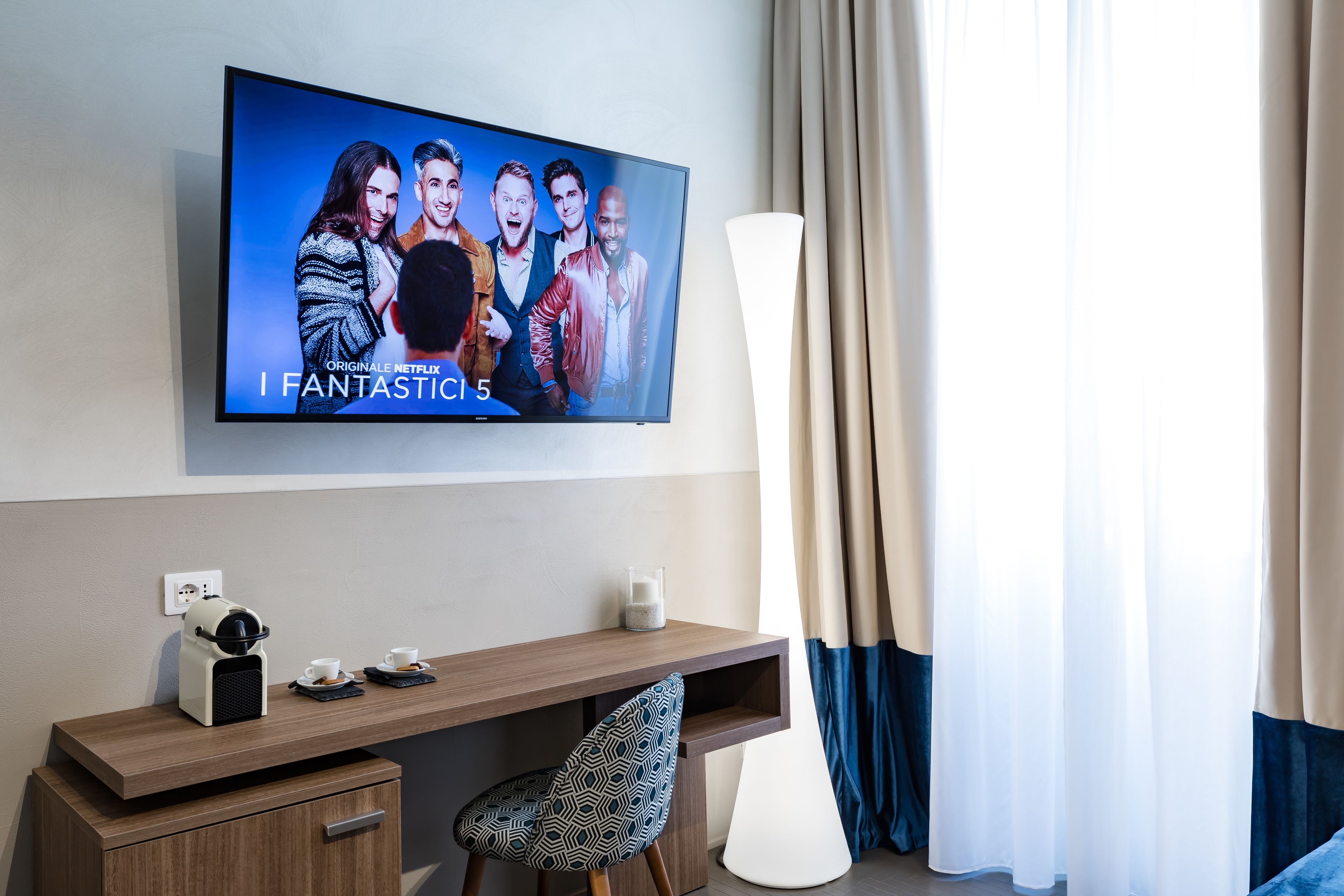 Ein modernes Hotelzimmer mit einem Flachbildfernseher, der den Film 'I Fantastici 5' zeigt, einem Holztisch mit Kaffeemaschine, einem gemusterten Sessel und einer großen Stehlampe.