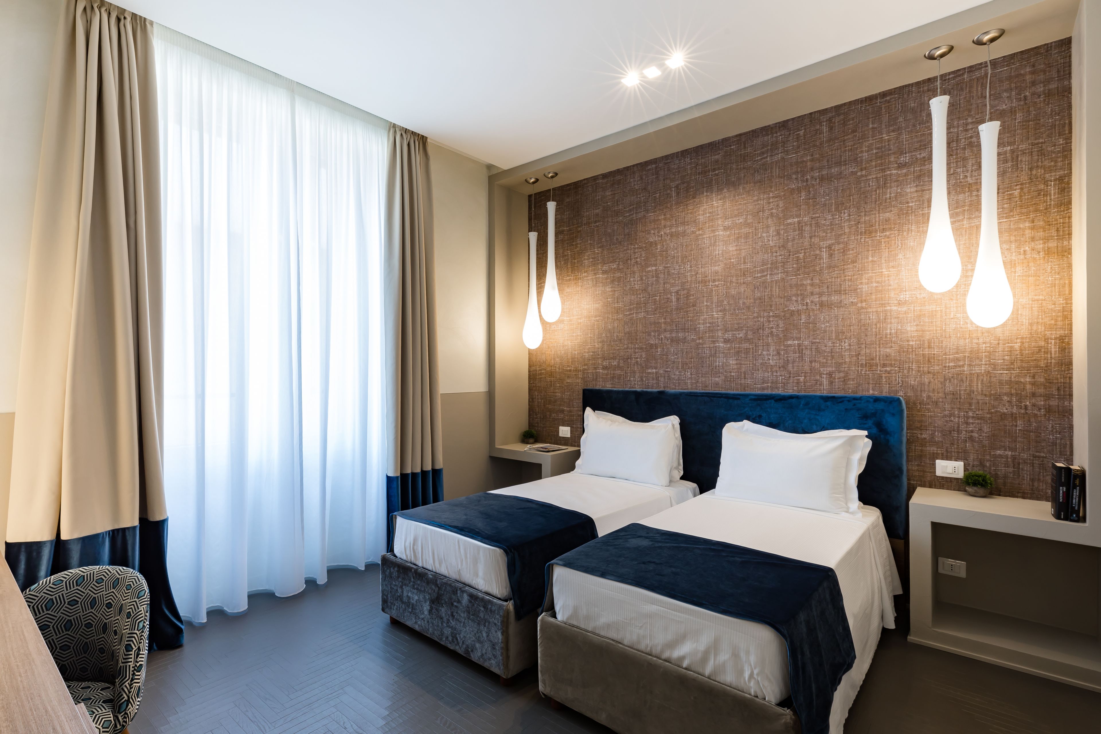 A hotel room with two beds, a large window with curtains, and modern pendant lights.