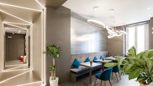 Un espace de restauration moderne avec un long couloir menant à un coin salon. On y trouve des tables et des chaises, ainsi qu'une grande plante au premier plan.