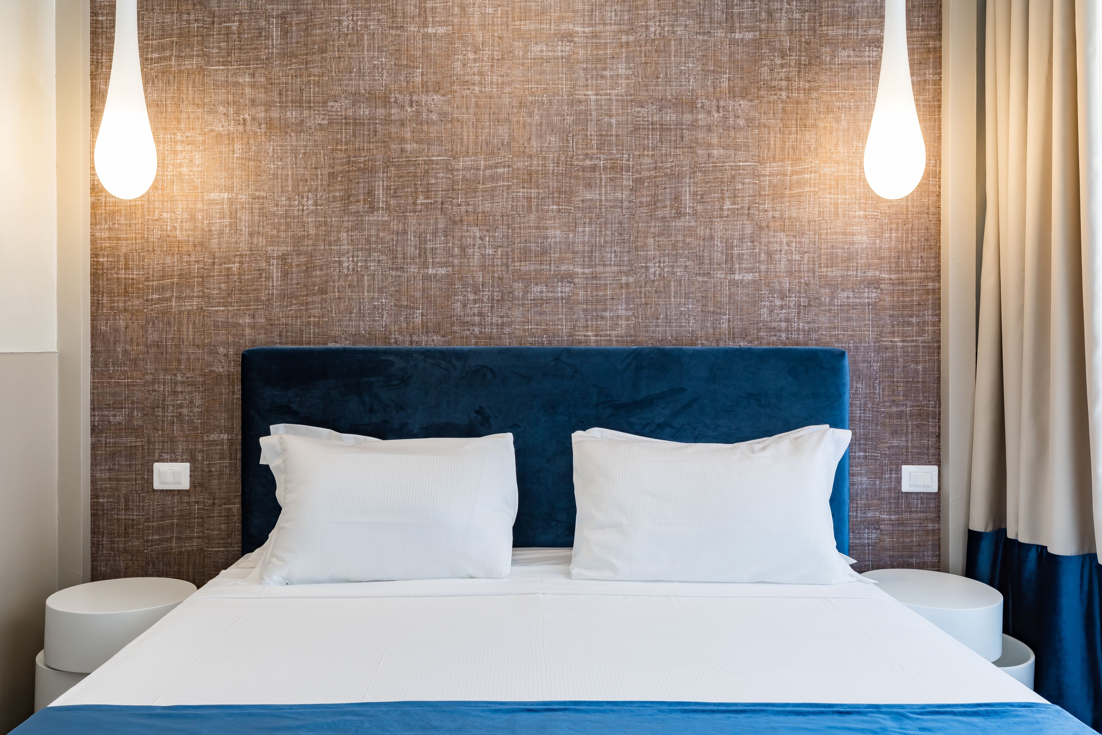A neatly made bed with white linens and a blue accent, a dark blue headboard, and two bedside lamps.