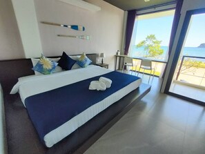A modern hotel room with a large bed, a view of the sea, and a balcony with a table and chairs.