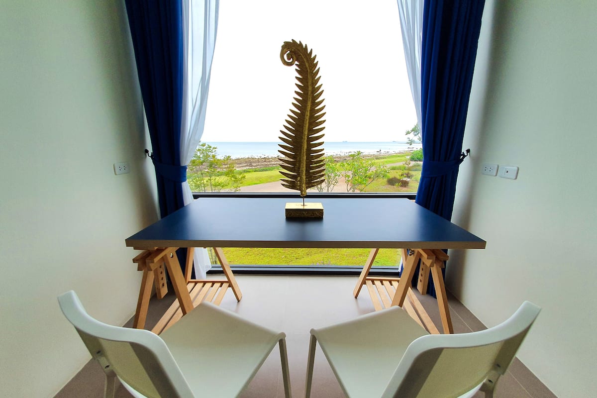 A room with a table and two chairs, a window with curtains, and a view of the beach.