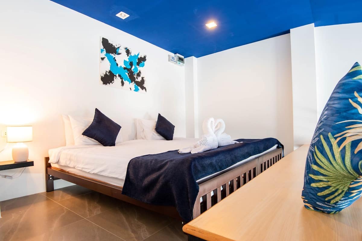 A hotel room with a bed, a bedside table, a lamp, a wall art piece, and a blue pillow.