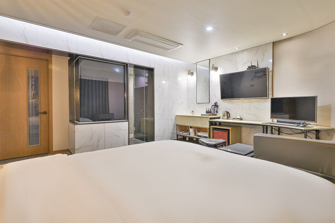 A modern hotel room with a large bed, a desk with a computer, a TV, and a bathroom with a glass shower enclosure.