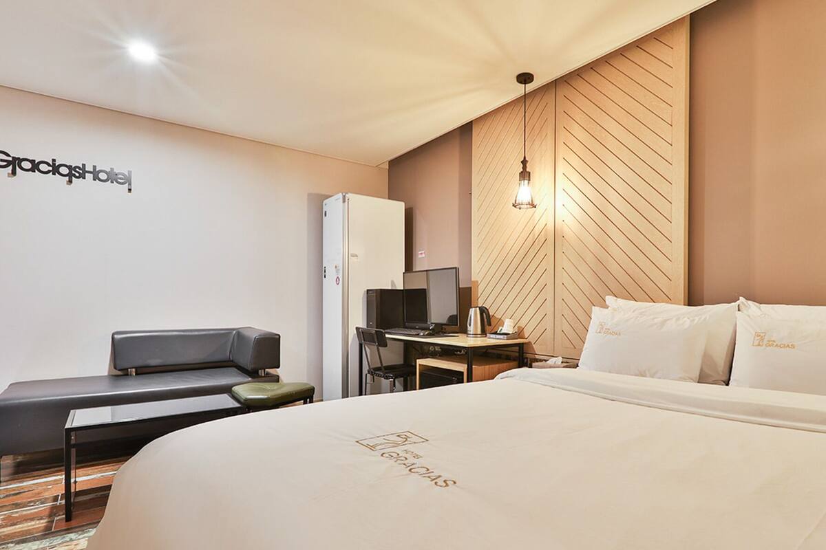 A hotel room with a bed, a desk, a chair, a refrigerator, and a wall-mounted light fixture.