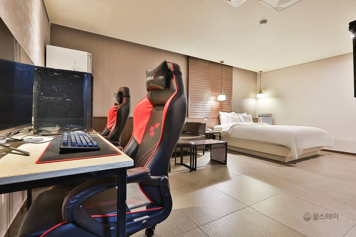 A hotel room with a desk, two gaming chairs, a bed, a bedside table, and a lamp.