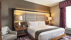 Ein Hotelzimmer mit einem großen Bett, zwei Nachttischlampen, einem Holznachttisch, einem Sessel und einem Fenster mit gemusterten Vorhängen.