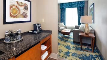 Habitación de hotel con tocador en el baño, cafetera y bandeja con botellas de agua y vasos. La zona de estar tiene un sofá, una mesa de centro y una lámpara.