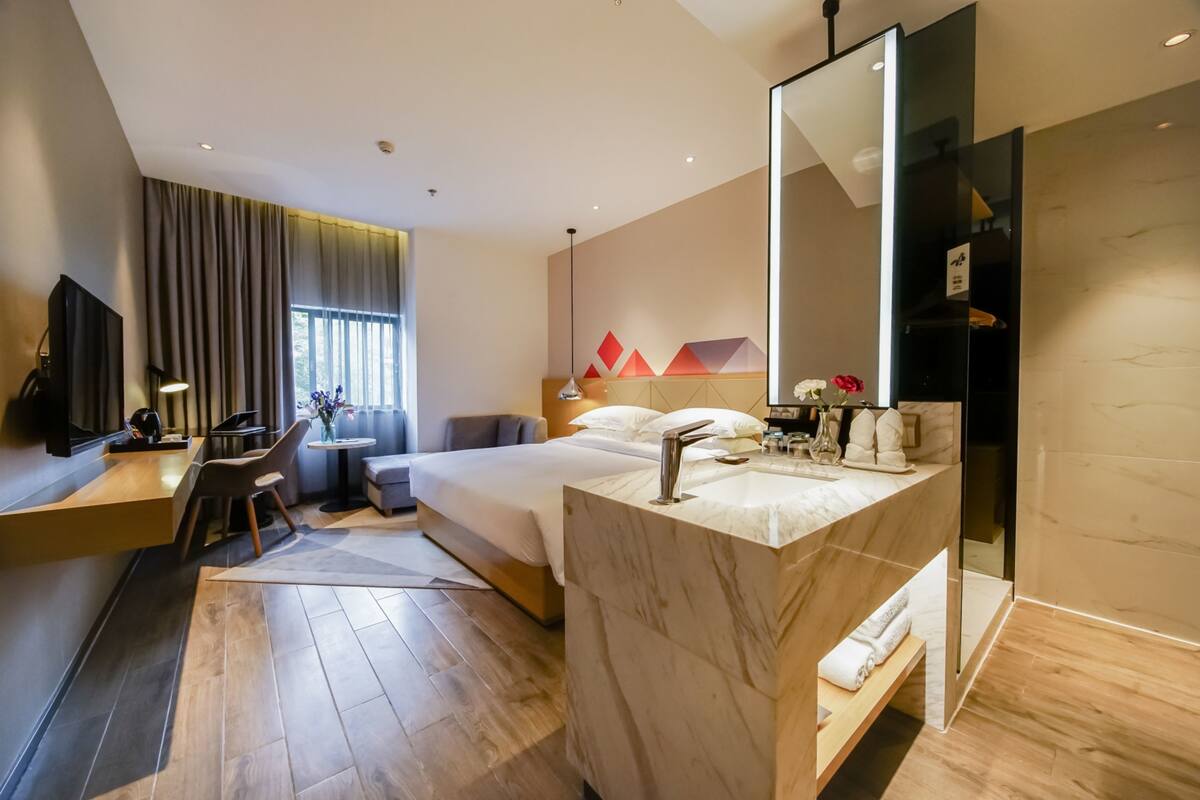 A modern hotel room with a large bed, a separate desk with a chair, a bathroom with a large mirror, and a TV mounted on the wall.